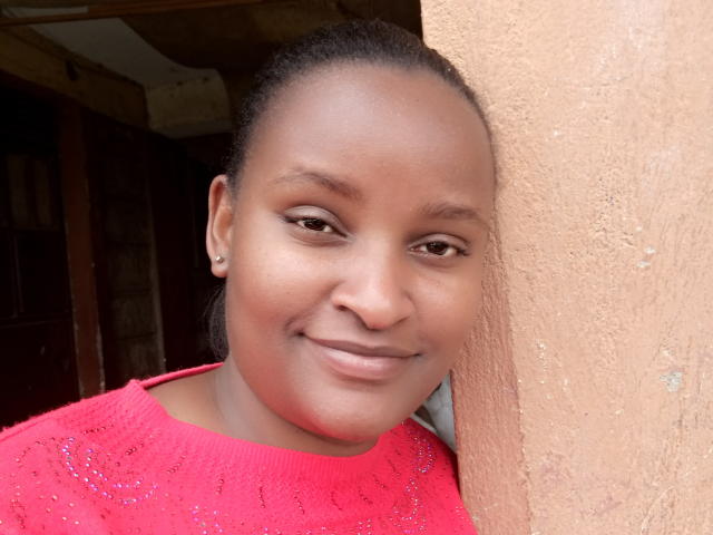 Close-up of Emphi69, a dark-skinned woman with a smooth, even complexion and subtle brown eyes. She has a slight, relaxed smile and wears a red, textured knit sweater with small sparkly details. Her hair is pulled back, and she has small, simple earrings. She leans against a rough, beige wall. The lighting is natural, highlighting her skin's smooth texture. The image focuses on her face, with a slightly off-center composition.