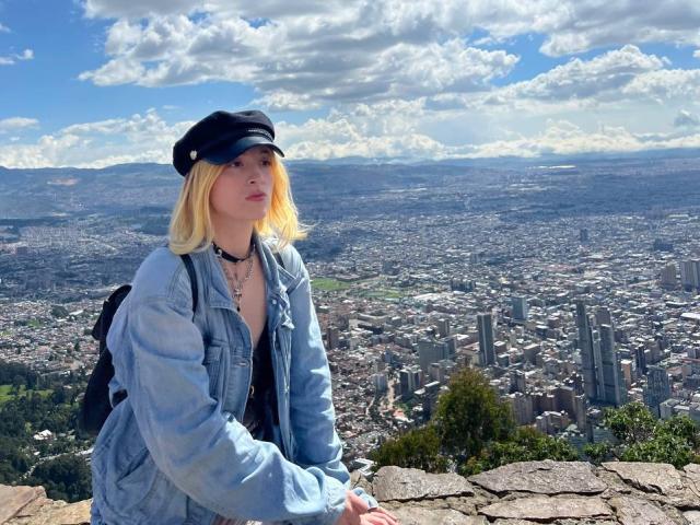 AshleySoy with blonde hair, black cap, denim jacket, and necklace, standing on rocky ledge, looking at city skyline, blue sky with fluffy clouds, and green patches in background.