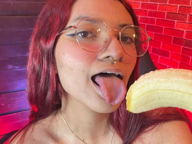 KiraRuby with red hair, glasses, and nose ring, tongue out, holding a banana near her face. She's wearing a gold chain and has a slight blush.