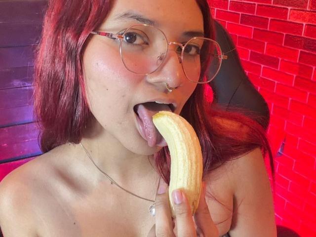 KiraRuby with red hair, glasses, nose ring, and tongue out, licking a banana, holding it with her right hand, showing bare shoulders, wearing a necklace.