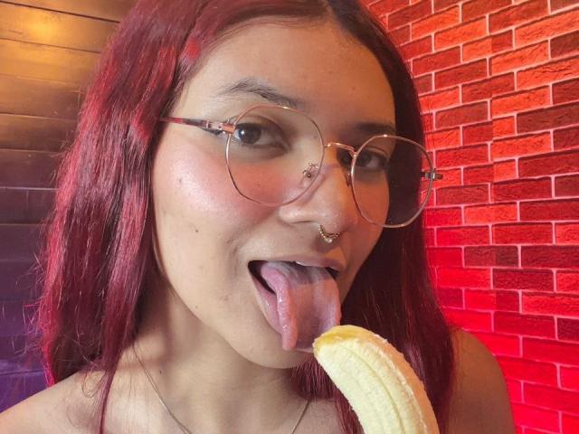 KiraRuby with red hair, glasses, nose piercings, and tongue out, licking a banana. She's got a cute face and a small nose ring on her left nostril.
