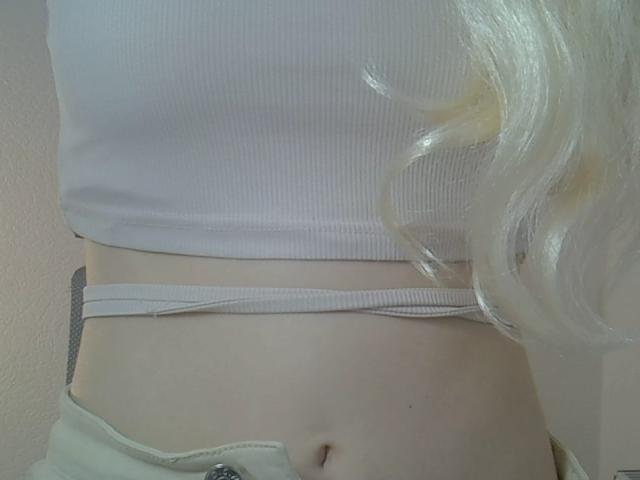 Close-up of DitaVira's midsection. She has a pale skin tone and wears a white ribbed crop top that exposes a small section of her lower abdomen. Her blonde hair, slightly wavy, cascades to the right. The crop top has a white elastic band that sits just below her chest. She also wears beige pants that are partially visible. The texture of the crop top is ribbed, contrasting with her smooth skin. The image is focused on her torso, with no other elements in the frame.
