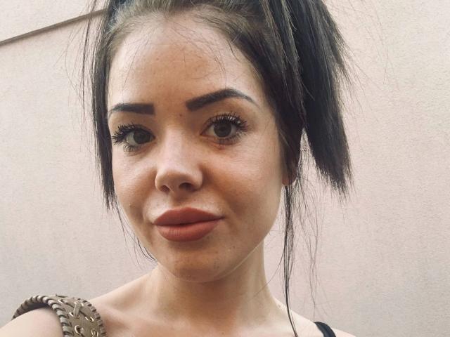 NicoleKatze-hot with dark hair in a messy ponytail, thick eyebrows, big brown eyes, and orange lipstick, wearing a spiked shoulder strap, smiling slightly at the camera.