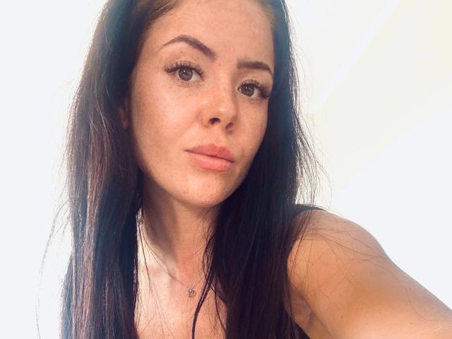 NicoleKatze-hot with long brown hair, freckled face, full lips, and thick eyelashes, wearing a black top, showing her bare shoulders, and a small necklace.