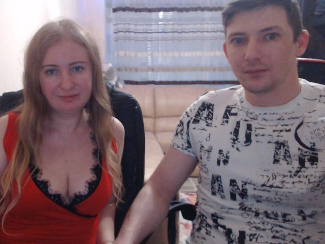 JenAndPeter com seios grandes, vestindo um vestido vermelho com renda preta, sentada ao lado de Peter, que tem cabelo castanho curto e está vestindo uma camiseta branca com estampa, em uma cadeira de escritório preta..
