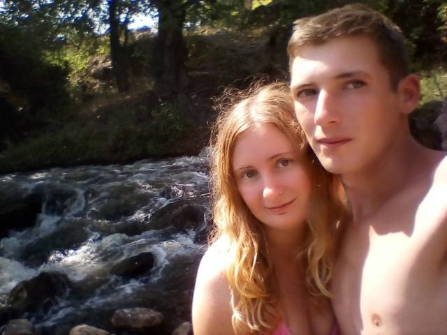 JenAndPeter, a blonde woman with wavy hair and small tits, stands shirtless next to a brown-haired man with a slim, muscular build, by a flowing creek.