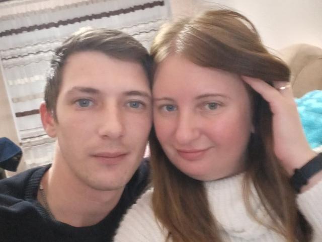 A close-up selfie of JenAndPeter. JenAndPeter, both with fair skin, are positioned in the foreground. Jen has short brown hair, light blue eyes, and a slight smile. He wears a black sweater. Peter has long red hair, blue eyes, and a gentle smile, holding his hair with his right hand. He wears a white turtleneck. The image has a casual, intimate feel. The composition centers on their faces, with soft lighting highlighting their features. A blue pillow is partially visible in the background.