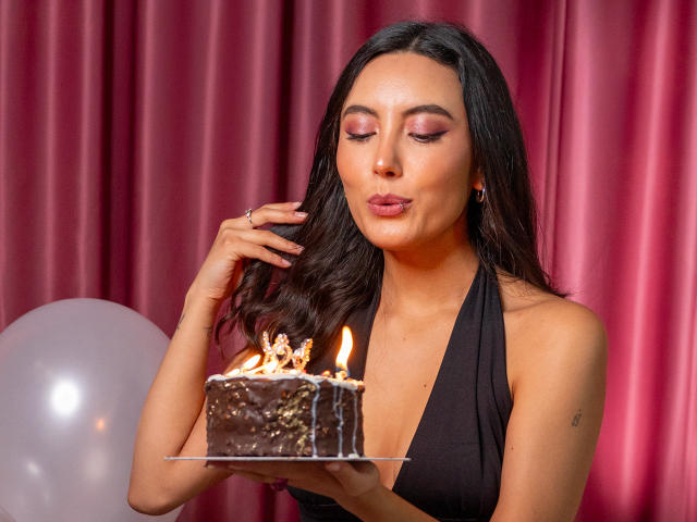 CarlaFranco, avec ses cheveux foncés, son fard à paupières rose et sa robe noire à encolure dégagée, souffle sur un gâteau au chocolat avec des bougies, tenant le gâteau devant elle, avec un fond de rideau rose et un ballon blanc..