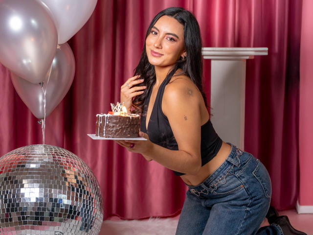 CarlaFranco, avec des cheveux noirs, une poitrine de taille moyenne, portant un haut noir à enfiler et un jean, tenant un gâteau au chocolat, debout près d'une boule à facettes et de ballons argentés..