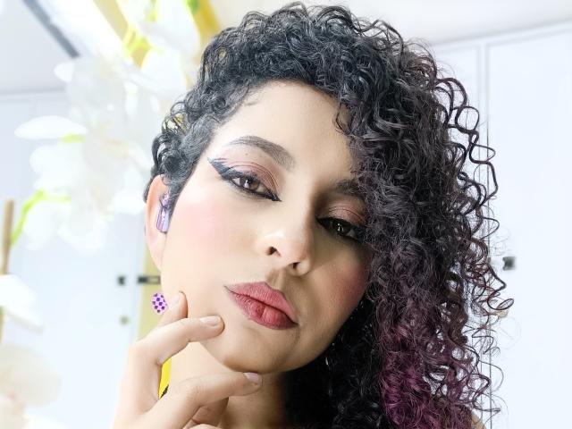 IrinaSweett has curly dark hair, pink eyeshadow, black eyeliner, and red lipstick. She's touching her chin with her finger, wearing purple earrings, and looking at the camera.