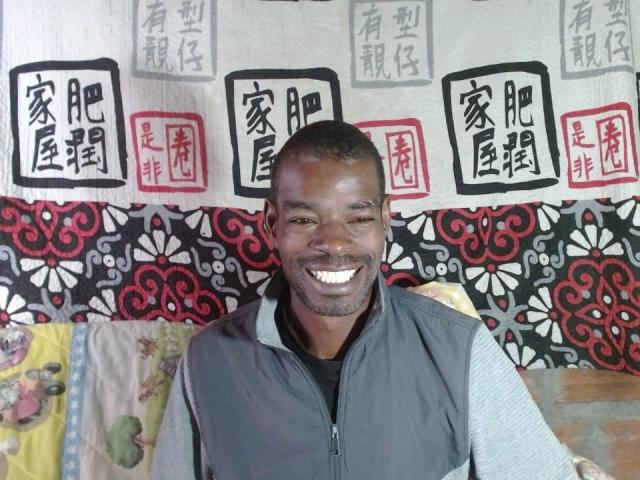 TaironTuck, a medium-skinned black man with a short haircut and a beard, smiles broadly. He wears a grey vest over a black shirt. The background features Asian-style signs with black and red characters on a white banner, and a patterned fabric with floral and geometric designs. His teeth are visible, and he has a slightly square jaw. The texture of his vest is smooth, contrasting with the fabric's intricate patterns.