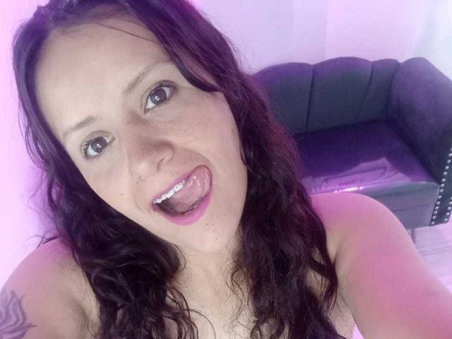 AriannaSeete has long, wavy dark hair, pale skin, and is sticking out her tongue with a playful expression, showing her white teeth. She has a tattoo on her right arm.