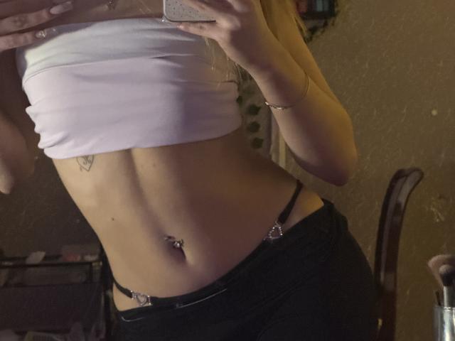 A close-up selfie of AmilySunray. She's wearing a white, form-fitting crop top that exposes her midriff, and black pants with a black thong visible. Her right hand holds a phone, partially covering her face. She has a small tattoo on her right side, below her ribcage. Her navel is pierced with a small, silver barbell. The lighting is dim, with a warm, brownish tone. Her skin is light, and she has a slender, toned physique.