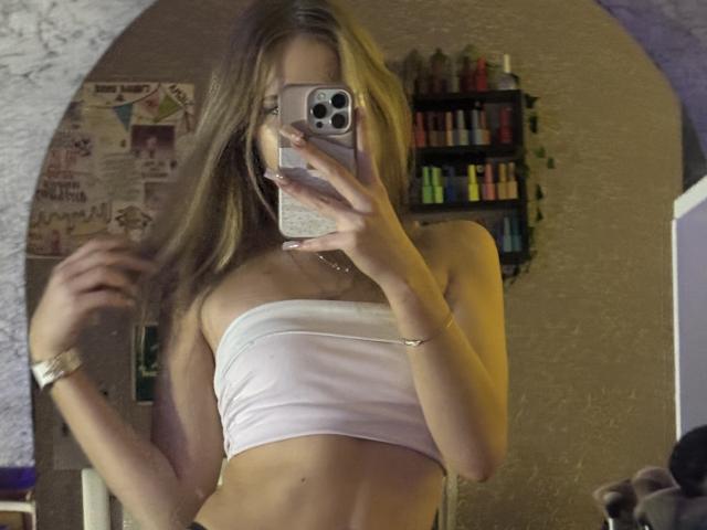 AmilySunray with long blonde hair, wearing a white strapless top, showing her bare tits in a mirror selfie, holding a phone with a glittery case.
