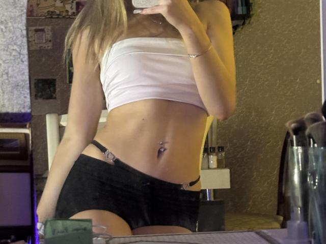 AmilySunray with blonde hair, white strapless top, black shorts, and belly button piercing, holding phone in mirror selfie, showing flat stomach and pierced navel.
