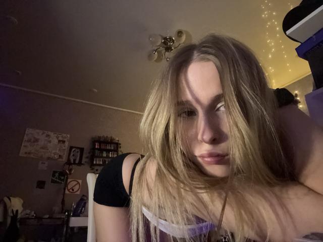 AmilySunray with blonde hair, slightly messy, leaning forward, eyes half-closed, wearing a black top, in a dimly lit room, soft light from fairy lights, ceiling fan above.