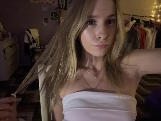 AmilySunray with blonde hair, wearing a white strapless top, holding her hair, looking at the camera, small tits, subtle necklace, dim room, faint yellow lights, serious expression.