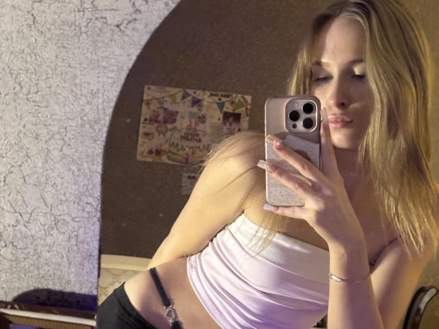 AmilySunray with blonde hair, pouty lips, wearing a white strapless top and black bottoms, taking a mirror selfie with her phone, showing her small tits and flat ass.