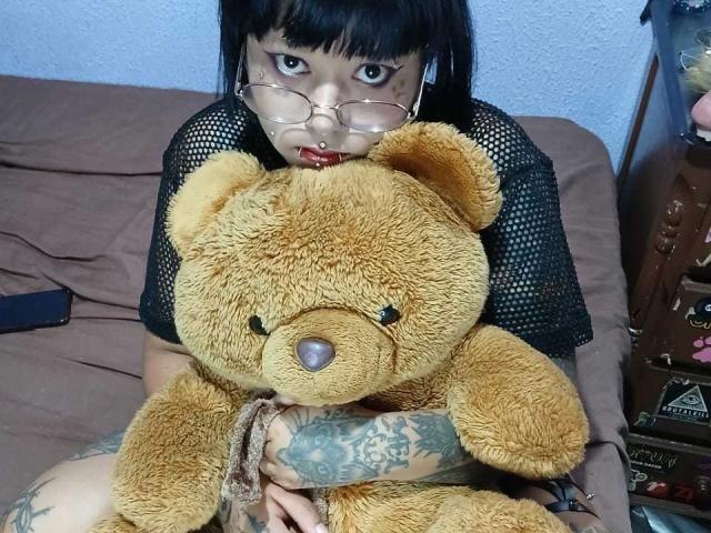 JenniferShaw with glasses, black mesh top, and tattoos, hugging a big, fluffy, brown teddy bear, sitting on a bed with brown sheets. She has black hair and red lipstick.