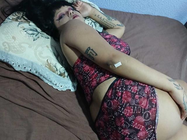 JenniferShaw lying on brown bed, wearing red and black patterned lingerie, showing her ass, tattoos on arm and side, bandage on arm, black hair, pillow with lace.