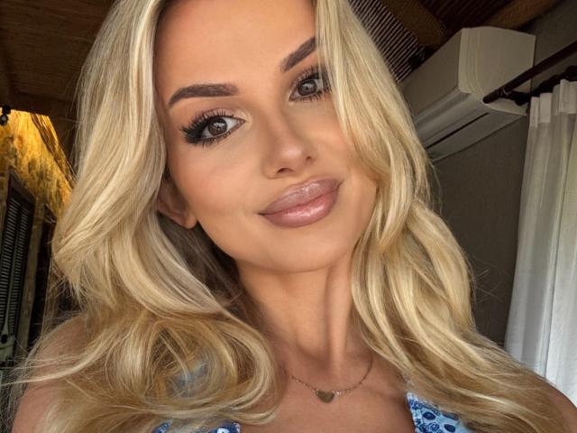 Blonde CorneliaDesire with wavy hair, full lips, and dark eyebrows, smiling at the camera. She's wearing a blue patterned top and a gold necklace.