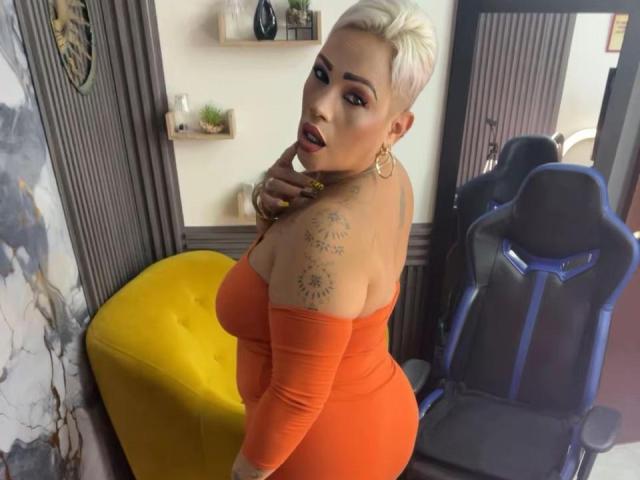 GianellaCox with short blonde hair, orange off-shoulder dress, gold earrings, finger near pink lips, tattoos on right shoulder, looking back, standing by yellow chair and blue gaming chair.