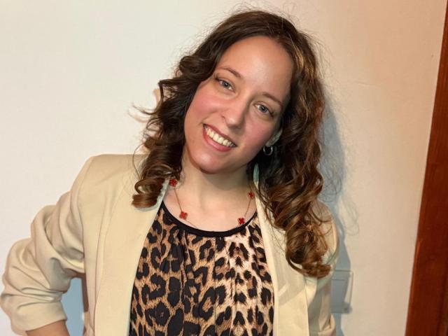 KirtyNella with curly brown hair, smiling, wearing a beige jacket over a leopard print top, red bead necklace, and earrings, standing against a plain wall.