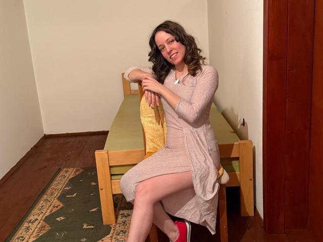 KirtyNella with curly brown hair, wearing a pink dress, sitting on a wooden bed with yellow pillow, smiling, hands on knees, red shoe, wooden floor, green carpet.
