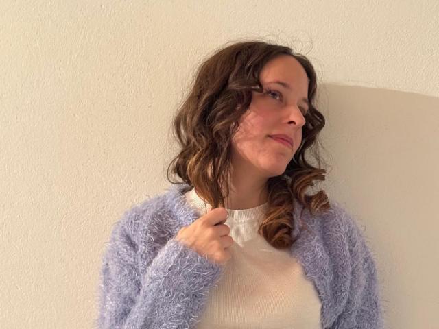 KirtyNella has wavy brown hair, a light purple fuzzy cardigan, and a white top. She's holding her hair with a slight smile, showing cute tits.
