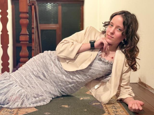 KirtyNella is lying on a carpeted staircase, wearing a beige blazer over a white, patterned dress. She has curly brown hair, a black wristwatch, and a relaxed expression.