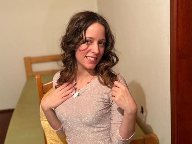 KirtyNella with wavy brown hair, smiling, wearing a sparkly pink long-sleeve top, hands near chest, showing small tits, standing in a room with wooden chairs and green table.