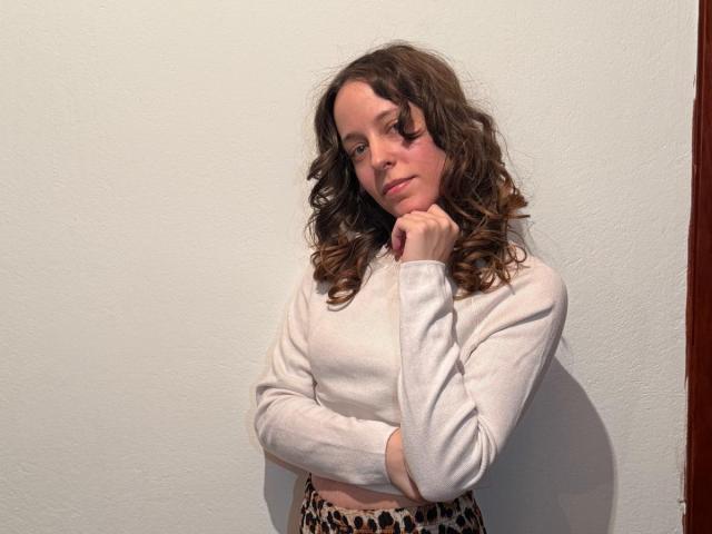 KirtyNella with curly brown hair, white long-sleeve sweater, and leopard-print pants, standing against a wall, hand under chin, looking slightly down, with a subtle, pensive expression.