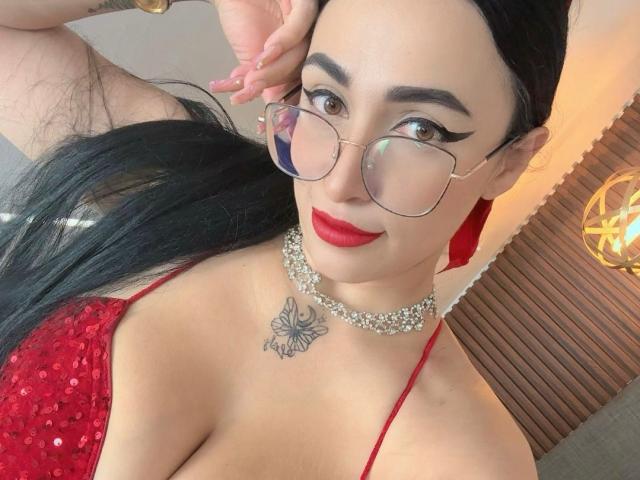 ScarlethSwann with glasses, red sequined top, pearl necklace, red lipstick, black hair, tattoo above boobs, hand up, smiling, brown eyes, clear skin, medium breasts.