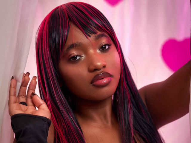 EmmaParisi with dark skin, pink and black hair, glossy pink lips, wearing a black fingerless glove, looking at the camera with a slight smile, pink heart in background.