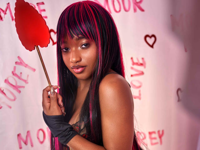 EmmaParisi with dark brown skin and pink-black hair, holding a red fan, wearing a black sleeve, looking at the camera with a slight smile, showing her small tits.