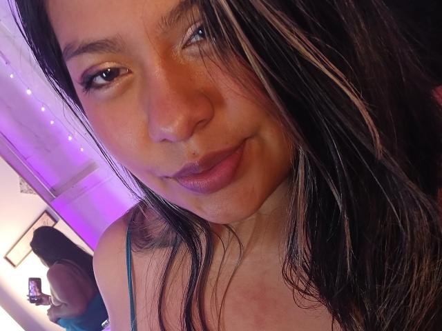 MelyOspina with dark brown, slightly wavy hair, and a subtle smile, wearing a blue spaghetti strap top, showing her soft brown skin and small nose.