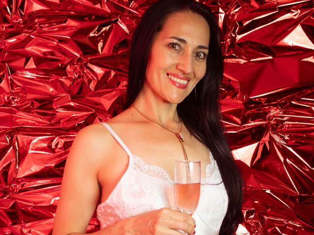 MiaMott has long black hair, fair skin, and is smiling at the camera. She's wearing a white lace camisole and holding a champagne glass. Her necklace has a red pendant. The background is crumpled red foil, creating a shiny, reflective texture. The lighting highlights her face and the glass. The image has a bright, festive feel.