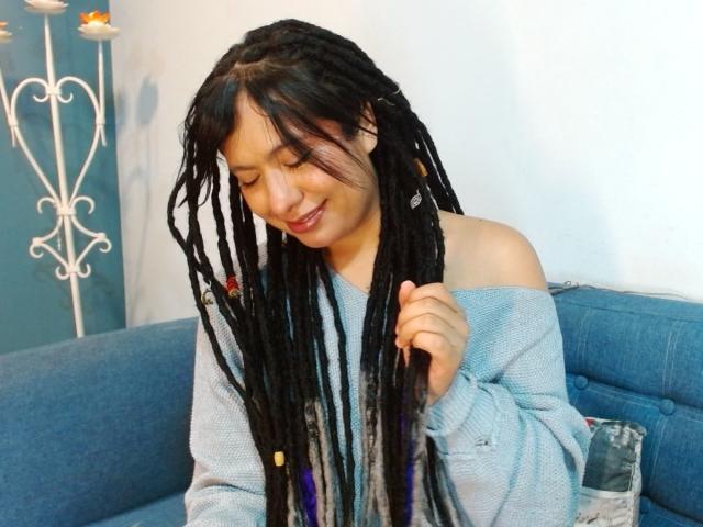 BeckyHuston with long black dreadlocks, wearing an off-shoulder light blue sweater, smiling, eyes closed, holding a lock of her hair, sitting on a blue couch.