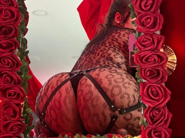 TessaMagic in lace lingerie, black strap harness, and red roses, showing off her big, round, shiny ass, looking back over her shoulder, with long dark hair.