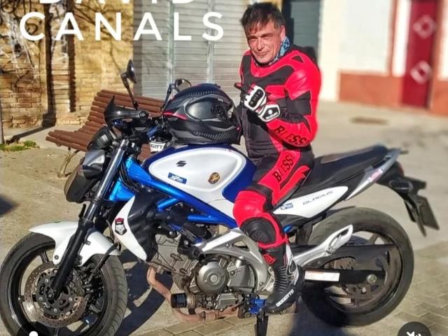 Dct3006, con un sedere prosperoso, indossa abbigliamento da motociclista rosso e nero, è seduta su una moto Honda blu e bianca, tiene in mano un casco e sorride alla luce del sole..