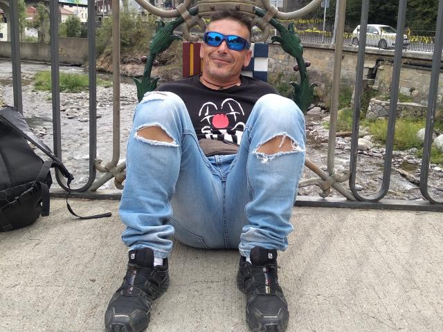 Dct3006, met een blauwe zonnebril, een zwart Spider-Man shirt, versleten jeans, zwarte laarzen, zittend op beton, lachend, met een zwarte rugzak aan de linkerkant..