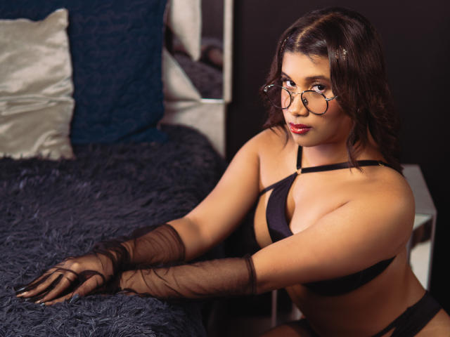 LoreDuval with glasses, black lingerie, and sheer gloves, sitting on a bed with dark blue, textured cover. She has brown hair, a curvy figure, and is looking at the camera.