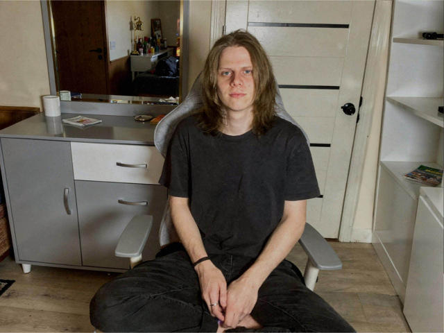 XSirHumpsX, with long brown hair, wearing a black t-shirt and black pants, sits cross-legged in a gray chair, looking straight at the camera, in a modest room.