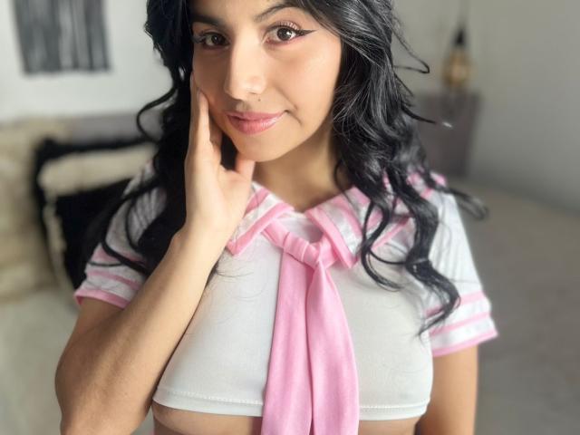 EmmaSollins with long black wavy hair, wearing a white and pink schoolgirl outfit, touching her cheek with a finger, smiling, showing her small pink nipples.