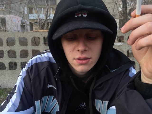XSirHumpsX, wearing a black hat and black and white jacket, holds a cigarette with a slightly open mouth, freckled face, and tired expression.