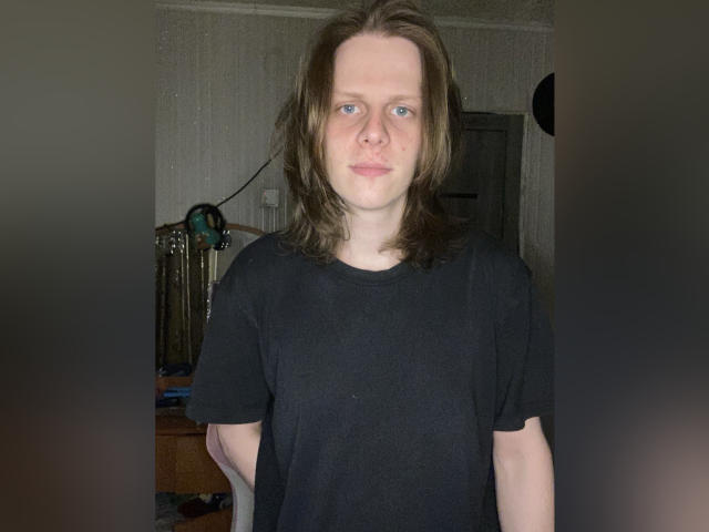 XSirHumpsX has pale skin, blue eyes, and medium-length brown hair, wearing a black shirt, standing with a neutral expression, hands behind back, in a dimly lit room.