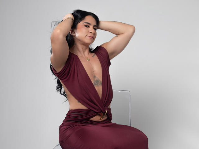 JuliethMaine with arms raised, wearing a deep burgundy dress, exposing her small tits and a tattoo between her breasts. She sits on a clear chair, smiling.