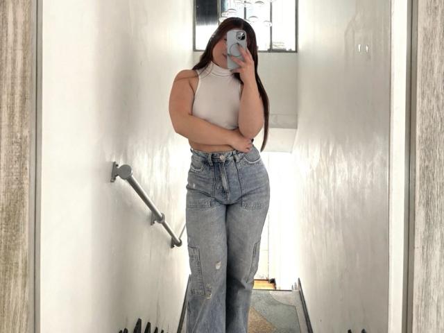 VicluX has long brown hair, a white sleeveless crop top, and high-waisted, blue, ripped jeans, holding a phone, covering her face, standing in a narrow white hallway.