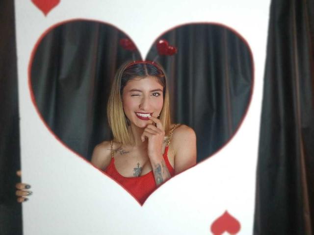 A photograph of Kaiix with shoulder-length blonde hair, wearing a red tank top, and a red headband with heart-shaped ears. Kaiix is smiling with one eye closed, biting her finger. She has tattoos on her chest and left arm. A heart-shaped cutout in a white frame partially covers her face. The background is dark. Kaiix's red headband has small heart decorations. Her nails are painted dark green. The frame has red heart symbols at the top left and bottom left.