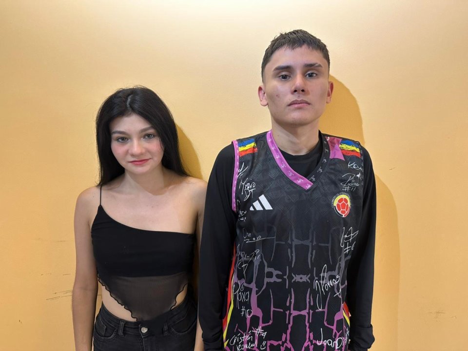Duo69sx con lunghi capelli neri, piccoli seni, indossa una maglia nera trasparente, è in piedi accanto a Duo69sx con capelli castani corti, che indossa una maglia da calcio nera e rosa, sfondo giallo..